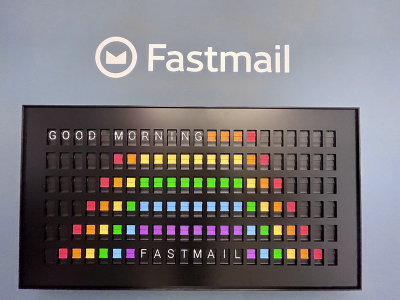 A split-flap board displaying 'Good morning, Fastmail,' overlaid on a rainbow